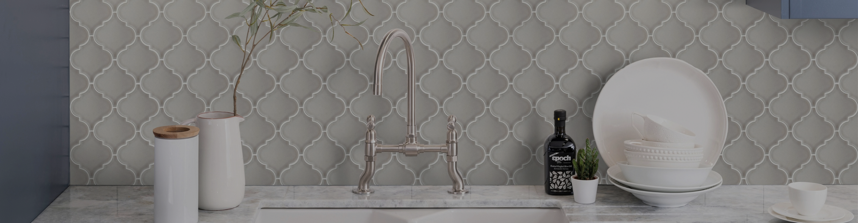 Bright kitchen countertop with a stainless sink and chrome faucet, white canisters with wooden lids, a small potted plant, and a large white bowl on a grey patterned backsplash.